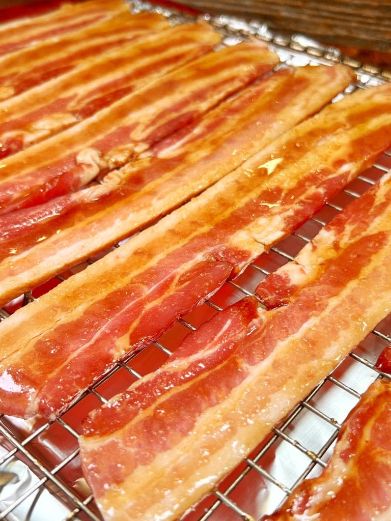 Close-up of several uncooked bacon strips arranged neatly on a wire rack, showcasing their marbled red and white patterns. The rack is placed on a shiny metallic surface, highlighting the fresh, glossy texture of the bacon.