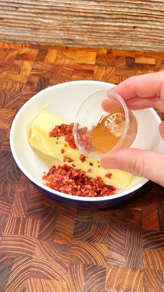 Hand pouring cinnamon from a small container into a bowl with a stick of butter and bacon bits on a wooden countertop to make candied bacon butter