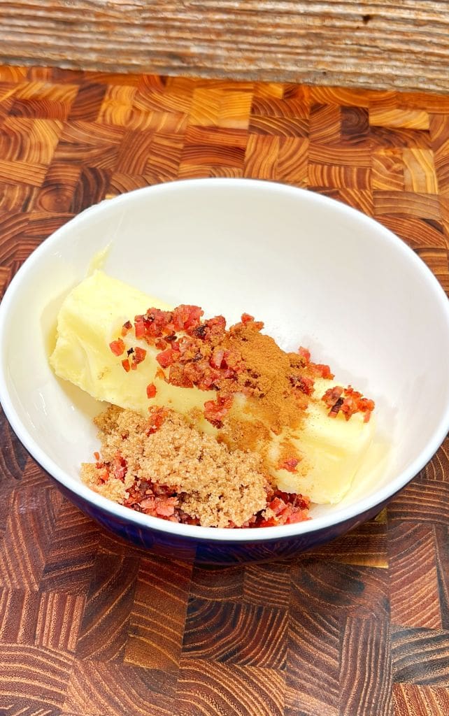 A white bowl is filled with ingredients on a wooden surface: a stick of butter, crumbled bacon, brown sugar, and a sprinkle of cinnamon.