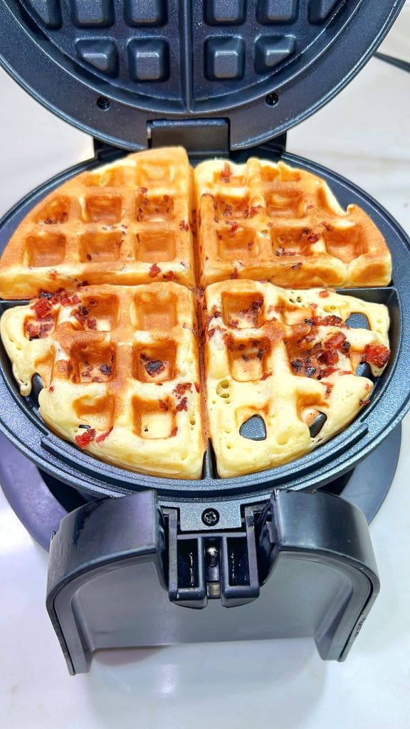 A waffle maker with a round, golden-brown bacon waffle inside. The waffle is divided into four sections and has pieces of bacon visible on the surface.