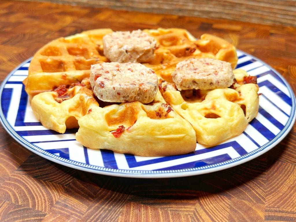 A plate of bacon waffles topped with chunks of herbed butter, served on a blue and white patterned dish, placed on a wooden surface.