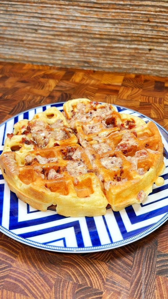 A golden-brown waffle with crispy edges and bits of bacon is served on a blue and white patterned plate, placed on a wooden table with a rustic backdrop.