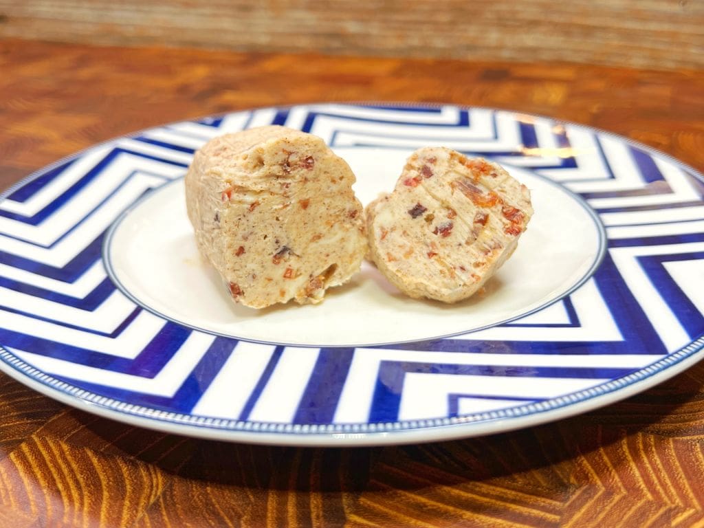 A close-up of two pieces of sliced cheese with visible herbs and spices, served on a decorative plate with a blue and white geometric pattern. The plate is placed on a wooden table surface.