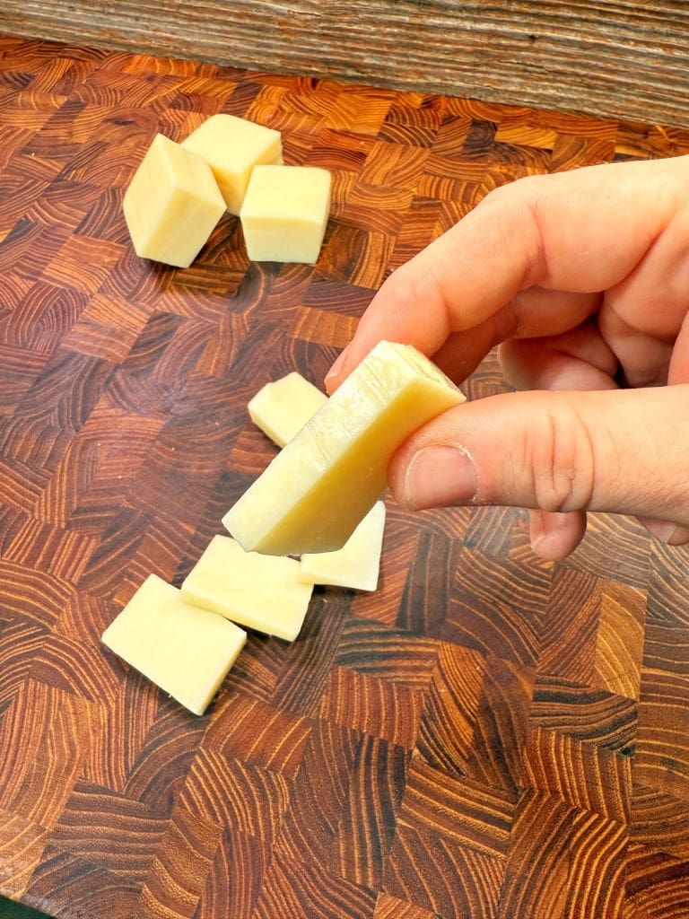 A hand holding a small cube of cheese over a wooden cutting board. Several other cheese cubes are scattered on the board. The wood has a rich, dark grain pattern.