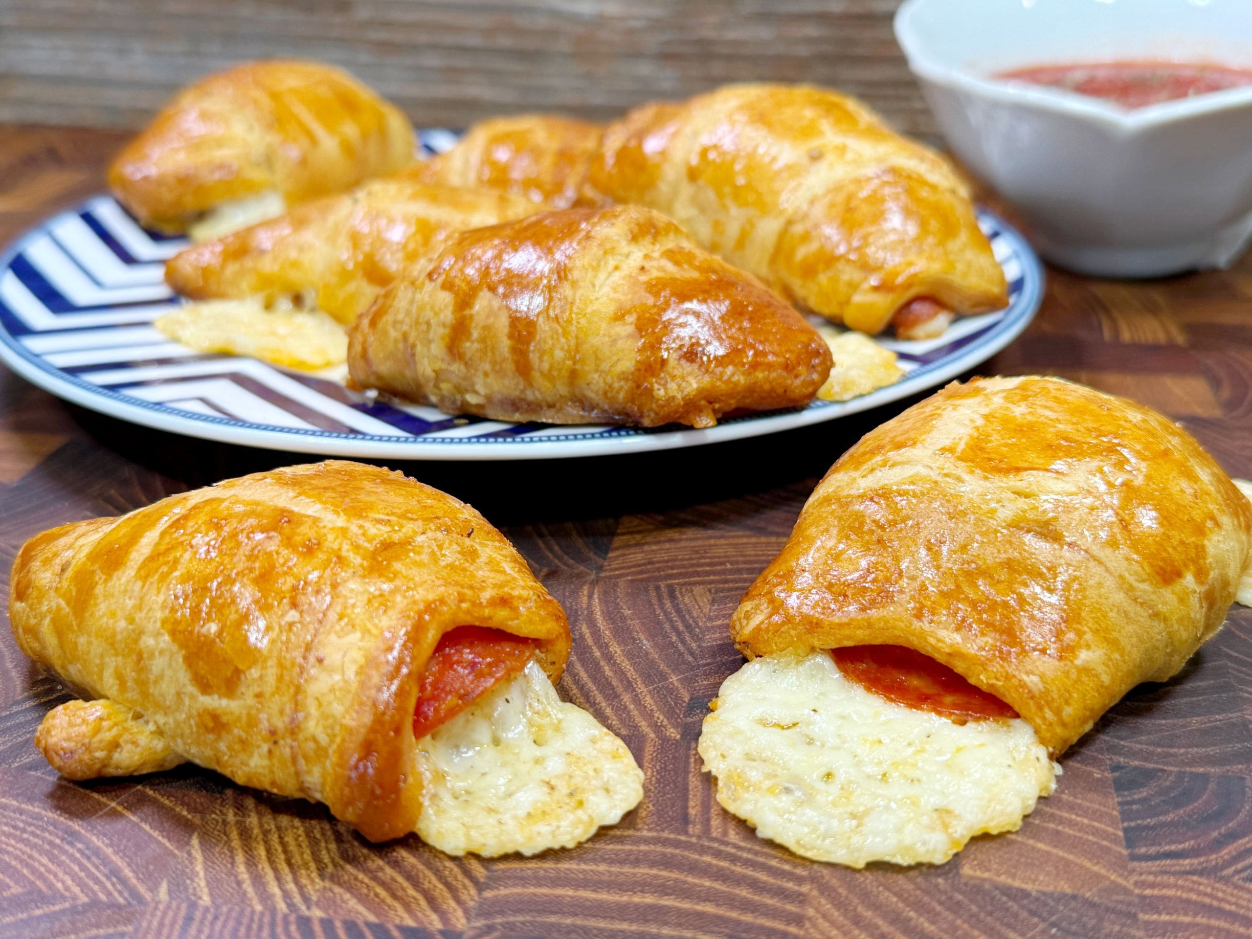 Golden-brown crescent rolls stuffed with pepperoni and melted cheese are displayed on a wooden surface. Some are on a blue and white patterned plate, with a small bowl of dipping sauce in the background.