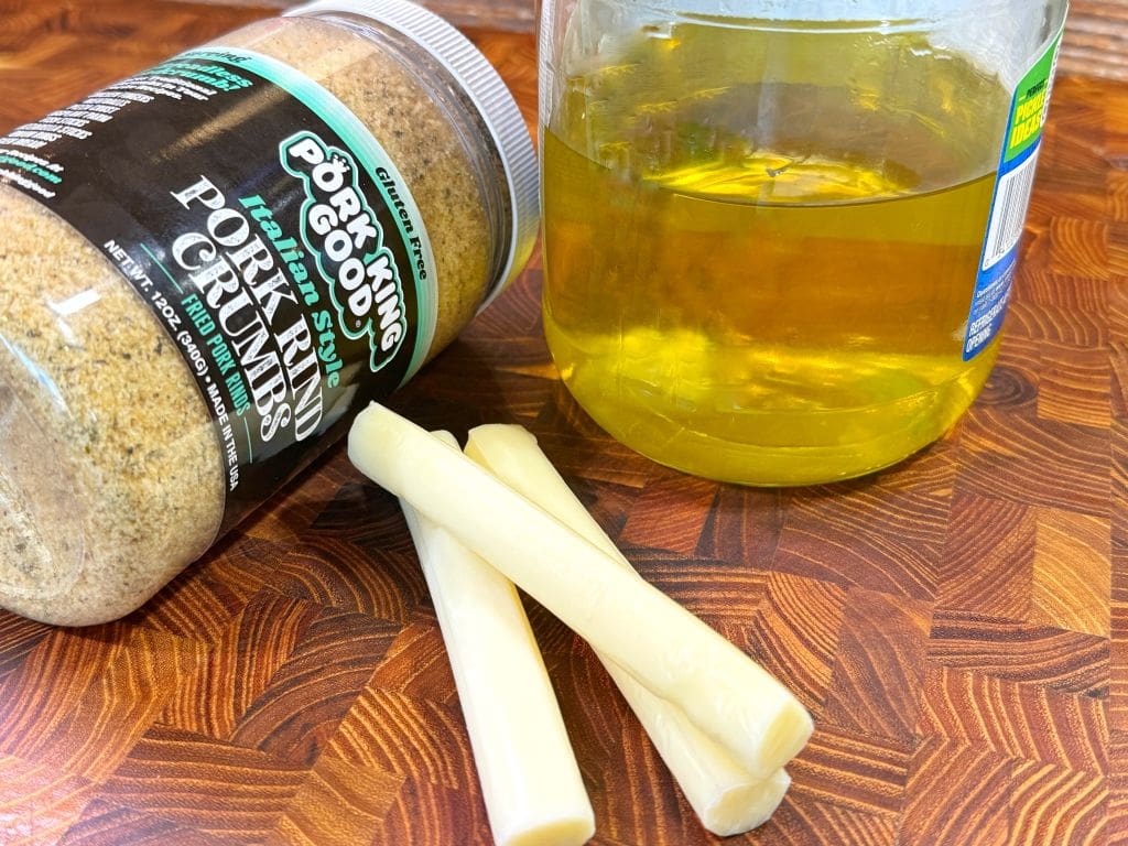 A container of pork rind crumbs, two string cheese sticks, and a jar of oil are on a wooden surface.