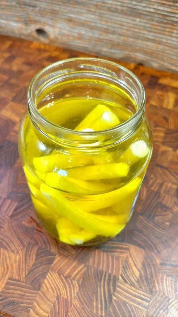A jar filled with yellow pickles submerged in brine, placed on a wooden surface with a rustic background. This will make the cheese have a fried pickle taste.