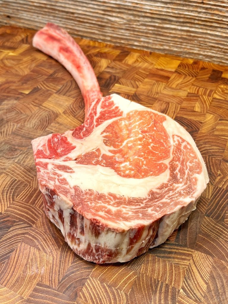 A raw tomahawk steak lies on a butcher block with visible marbling and a long bone handle. The wooden surface features a rich, patterned texture.