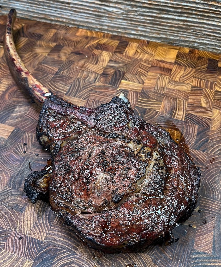 A reverse seared tomahawk steak rests on a wooden cutting board with a distinct marbled surface. The steak appears charred with visible juices, and the long bone is prominently extending from one side.