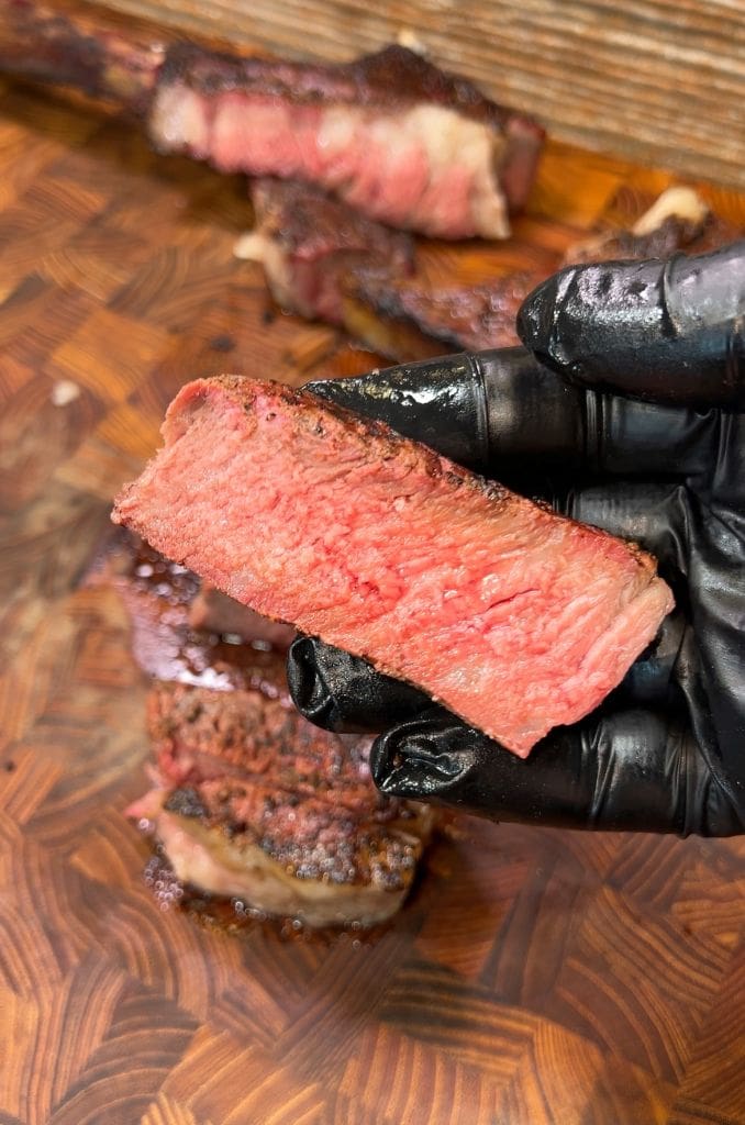 A gloved hand holds a juicy, medium-rare slice of smoked and reverse seared tomahawk steak over a wooden cutting board, with more slices in the background. The meat has a seared outer layer and a pink interior, highlighting its tenderness and doneness.