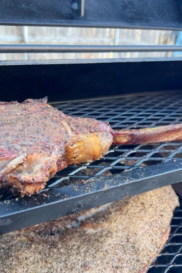 A seasoned tomahawk steak is being smoked on a grill, resting on a metal rack above other pieces of meat. The grill is open, revealing the charred, smoky texture of the steak. Nearby, a metal tray holds additional food items.