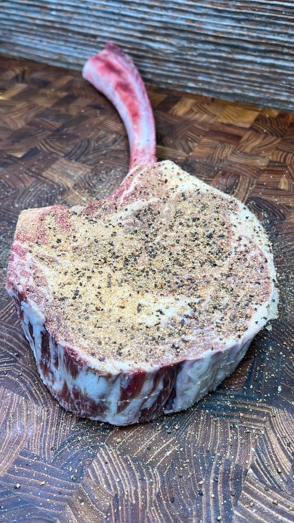 A raw, seasoned tomahawk steak rests on a wooden cutting board. The steak has visible marbling and a long rib bone, with seasoning sprinkled on top.