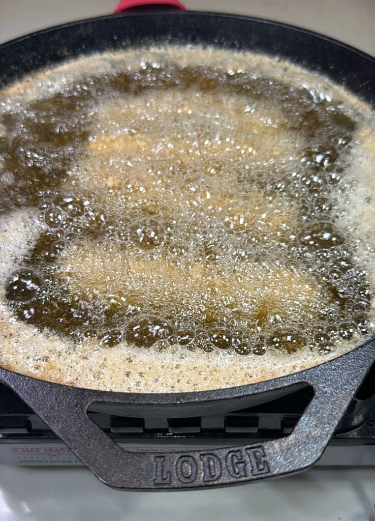 A Lodge cast iron skillet filled with bubbling oil is being used for deep frying. The oil is foaming at the surface, indicating something is being cooked inside it. The words Lodge are embossed on the skillet handle.