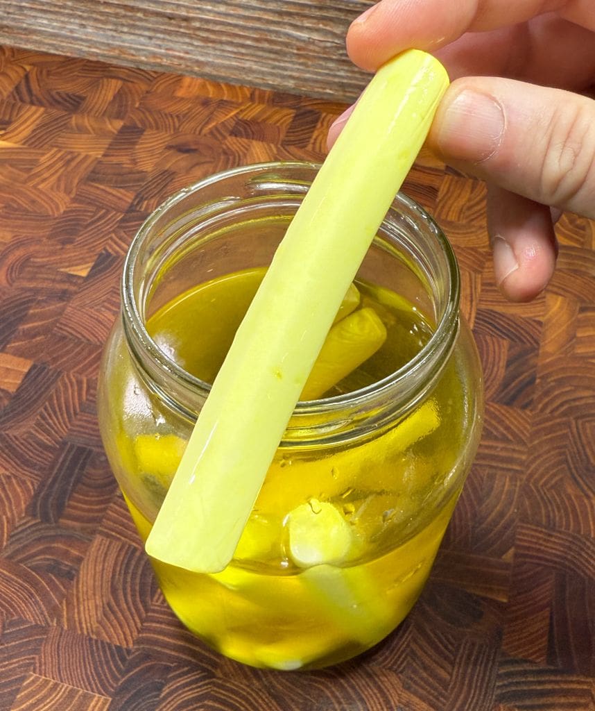 A hand holds a pickled cucumber spear above an open jar filled with more pickles and brine, set on a wooden surface. The pickles are bright yellow-green in color.