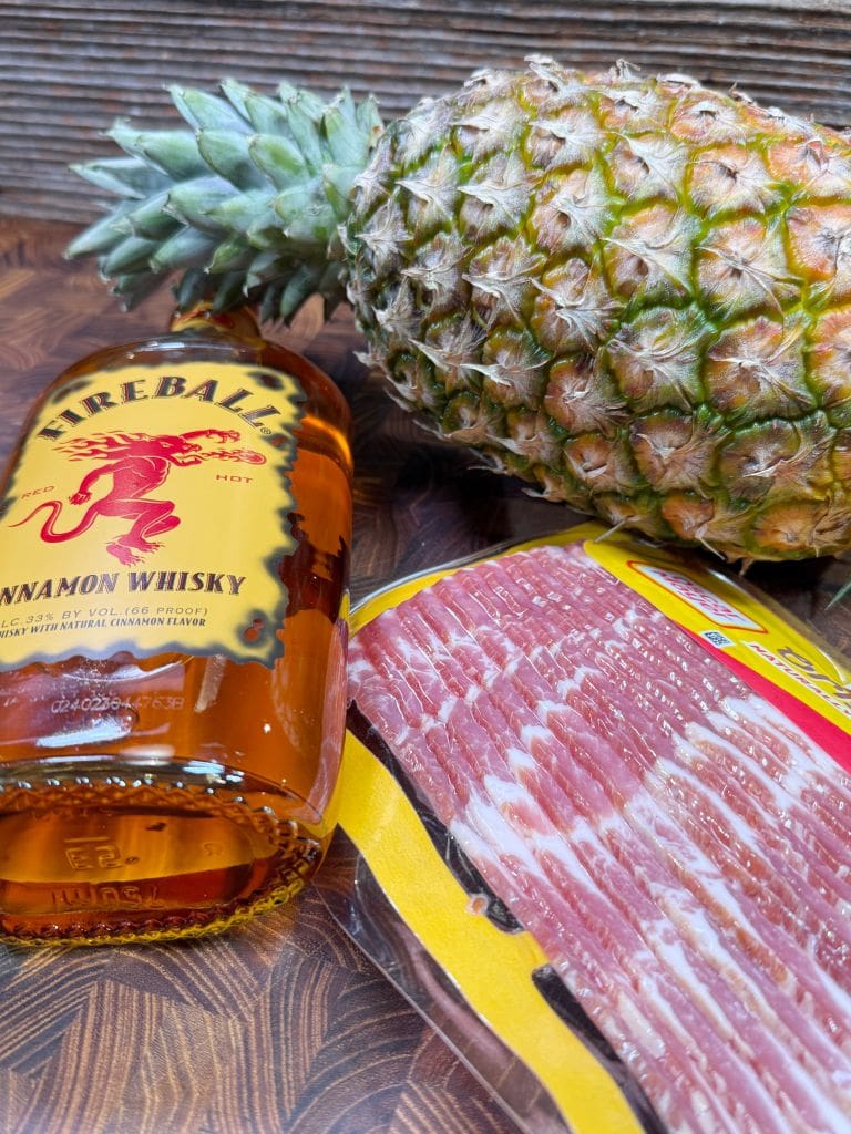 A bottle of Fireball Cinnamon Whisky lies on a wooden surface next to a fresh pineapple and a package of bacon.