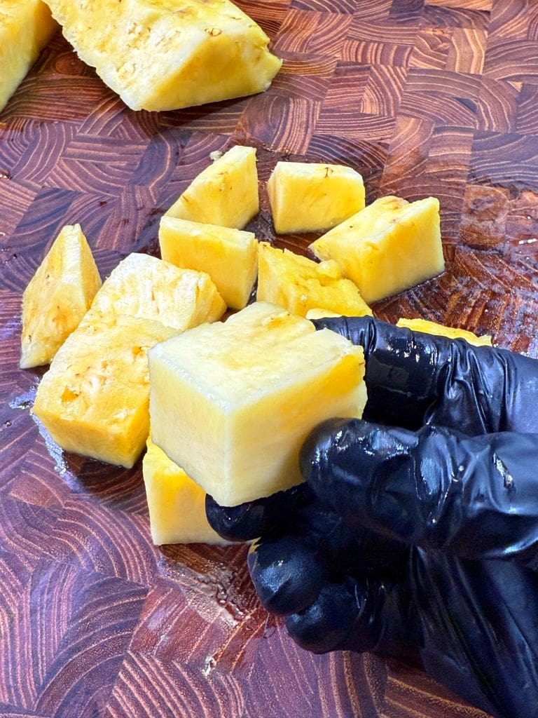 A hand in a black glove holds a cube of fresh pineapple above a wooden cutting board. Additional pieces of pineapple are scattered on the board, showcasing a yellow, juicy texture.