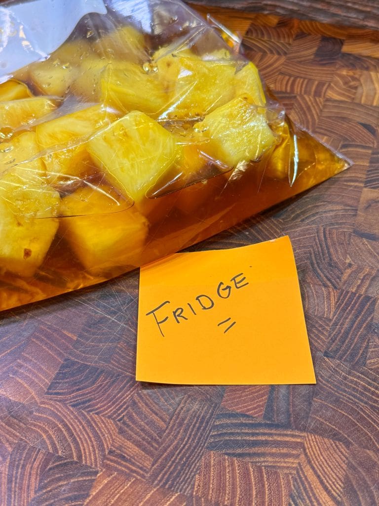 A plastic bag filled with pineapple chunks soaking in a liquid rests on a wooden surface. An orange sticky note nearby has the word FRIDGE written on it.