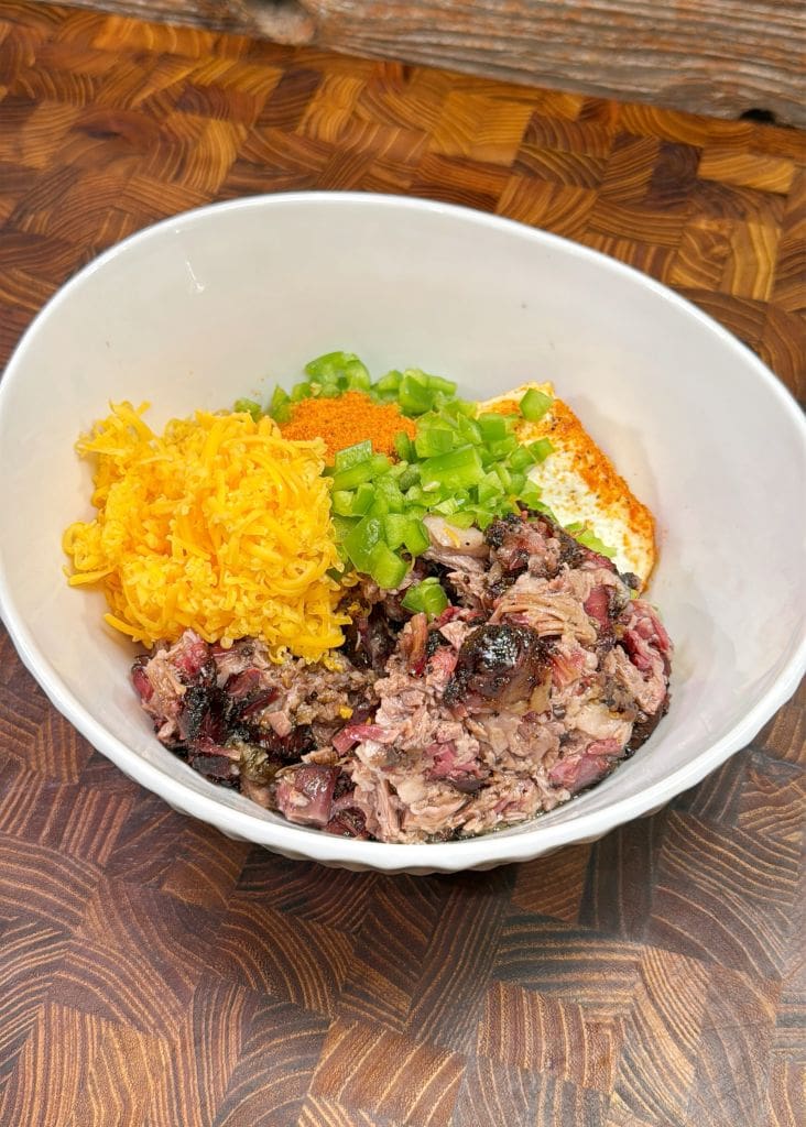 A white bowl containing shredded meat, grated cheese, green peppers, and a dollop of sauce on a textured wooden surface. The dish features a variety of colors and textures, highlighting the fresh ingredients.