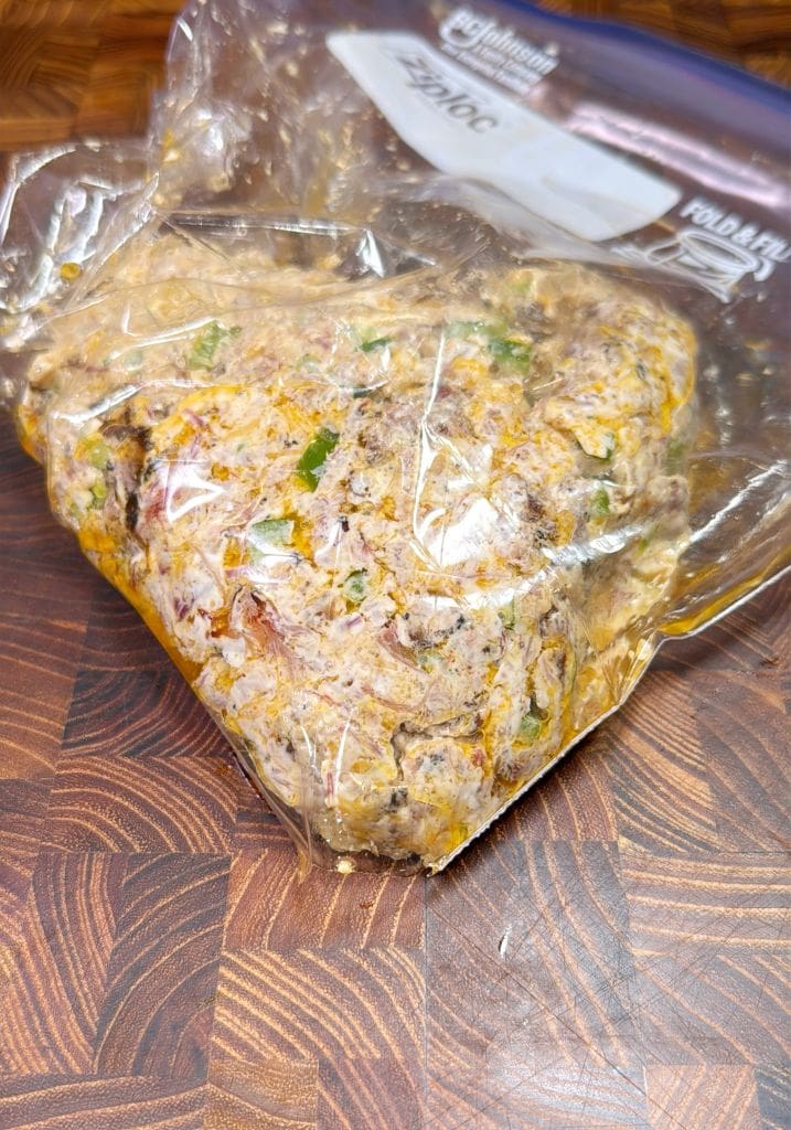 A clear plastic bag filled with a mixture of ground meat, herbs, and spices sits on a wooden countertop. The bags top is open, and the contents are visibly mixed with green and yellow flecks.