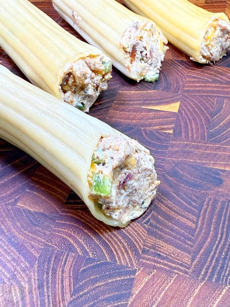 Close-up of uncooked manicotti pasta tubes stuffed with a creamy mixture of minced vegetables and possibly meat or cheese, placed on a textured wooden surface.