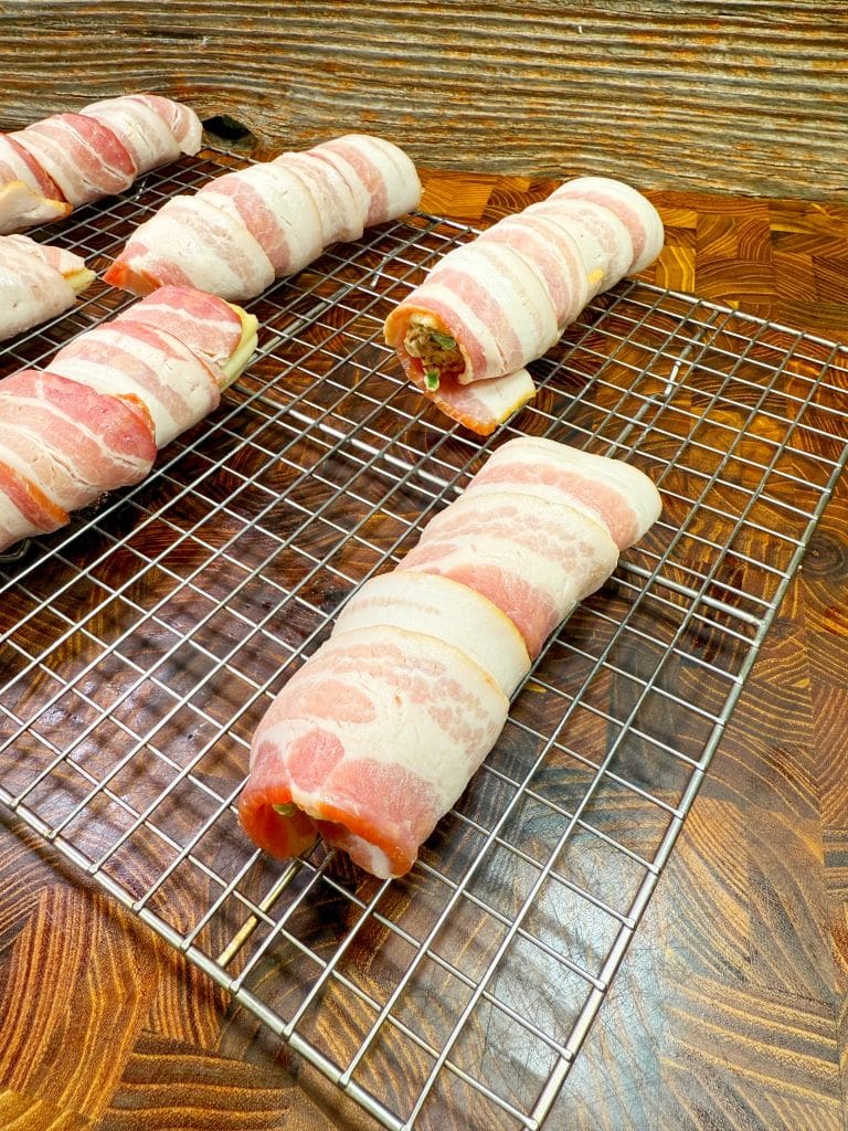 Bacon-wrapped stuffed chicken breasts rest on a wire rack placed on a wooden countertop. The bacon is neatly wrapped around each piece, and they are evenly spaced, ready for cooking.