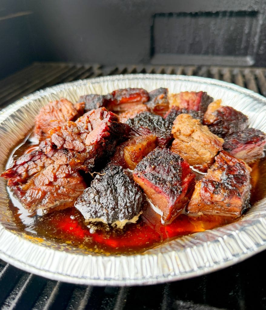 Close-up of a foil pan filled with charred, a juicy beef brisket burnt ends recipe, resting on a grill. The meat appears caramelized with a glossy finish, surrounded by a rich, savory sauce.