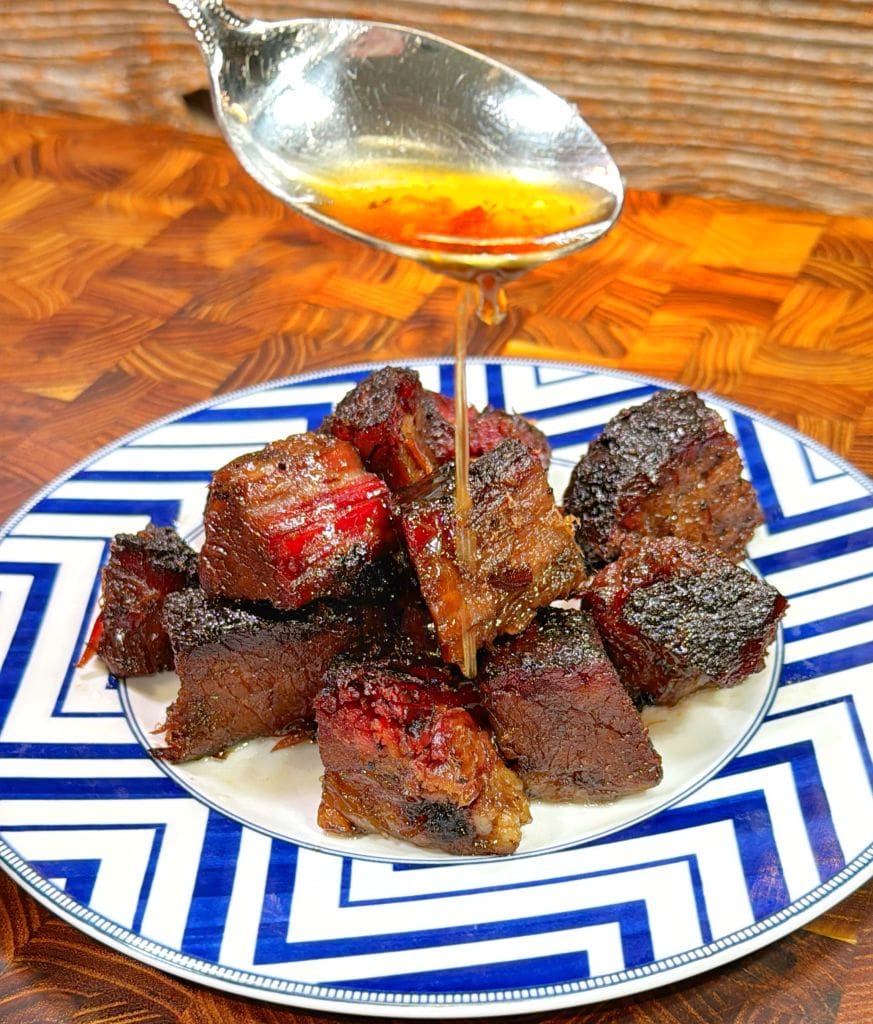 A spoon drizzles honey over a plate of grilled beef cubes on a blue and white patterned plate, set against a wooden background.