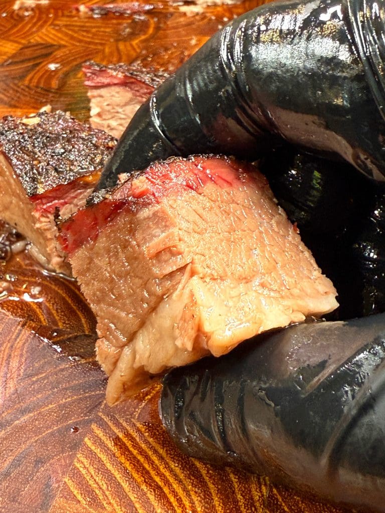 A close-up of a gloved hand holding a thick slice of juicy, cooked candied brisket with a dark, caramelized crust on a wooden cutting board, showcasing its tender and succulent texture.