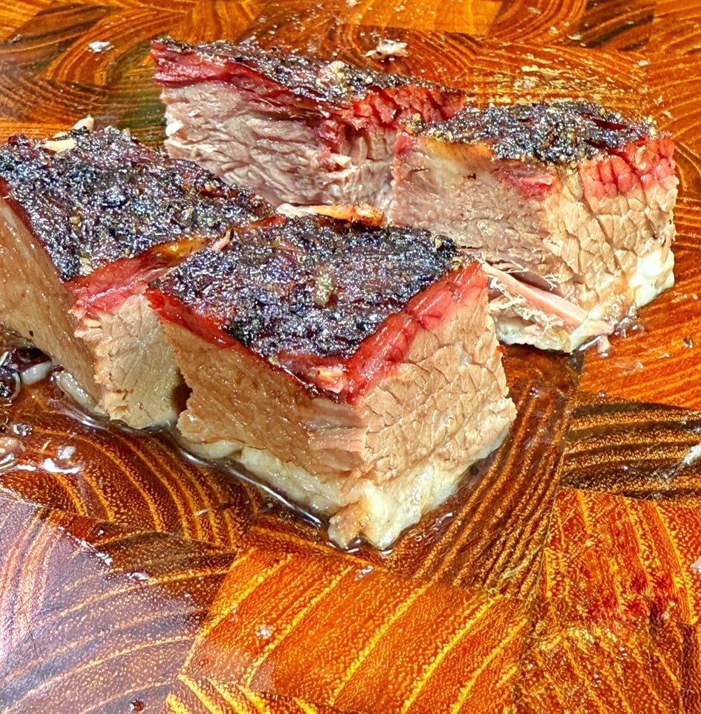 Close-up of sliced, juicy brisket with a crispy, charred exterior on a wooden cutting board. The meat appears tender, showcasing candied brisket burnt ends with visible layers of bark and a mesmerizing smoke ring.