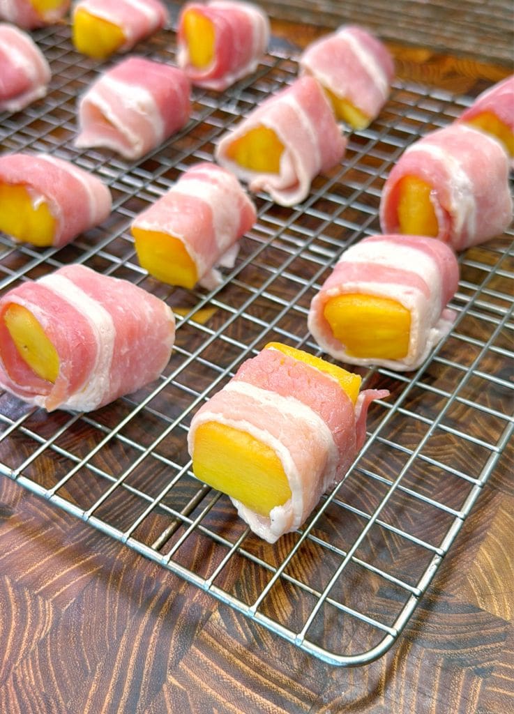 Bacon wrapped pineapple slices of fruit, resembling mango or similar, placed on a wire rack over a wooden surface.