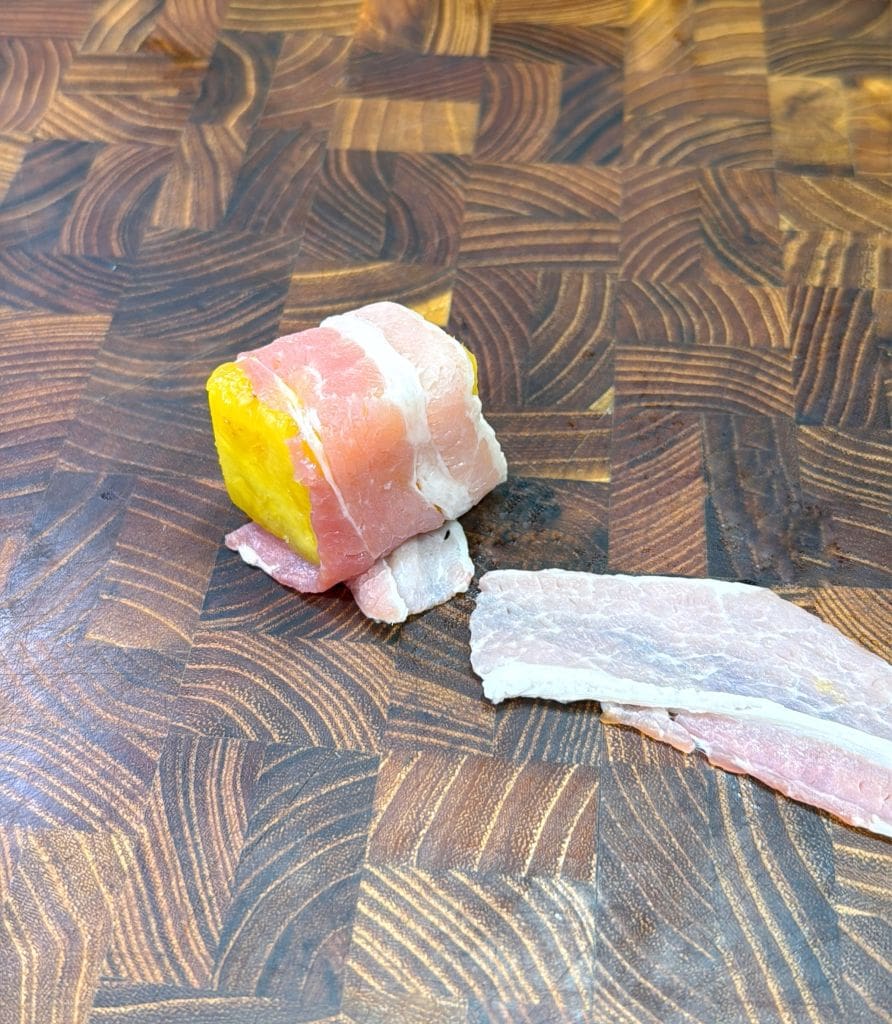 A cube of yellow squash wrapped in raw bacon sits on a wooden cutting board with intricate patterns, with an additional strip of bacon lying next to it.