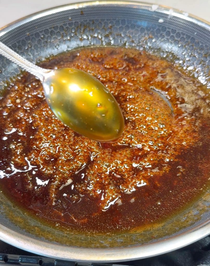 A metal spoon scooping caramelized sugar from a frying pan. The sugar mixture is brown and bubbling, with a glossy appearance. The spoon reflects some light, adding a golden hue to the scene.