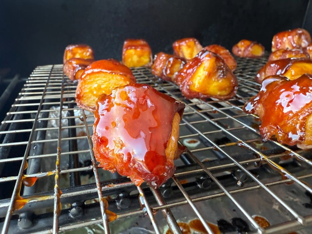 Close-up of glazed meat bites on a grill rack, with a shiny sauce coating. The grill is positioned over a drip pan, capturing excess juices. Several pieces are seen in the background, evenly spaced and caramelized.