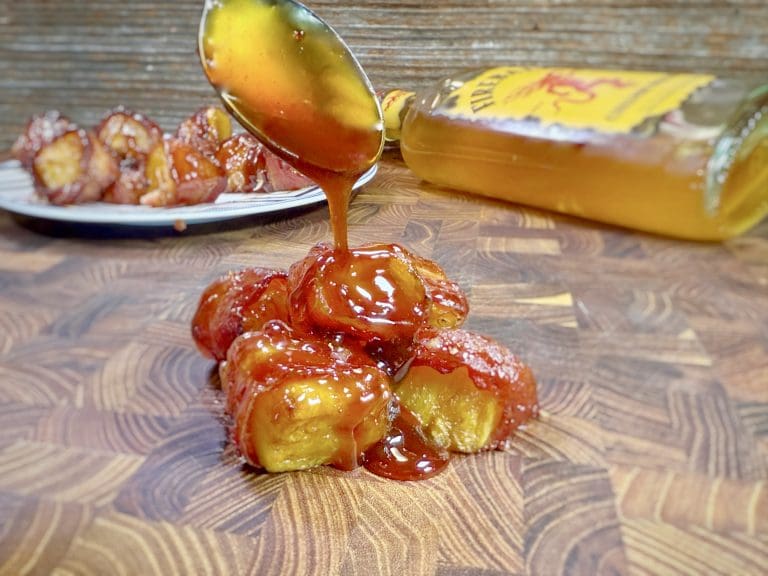 A spoon drizzles caramel sauce over a stack of sliced bananas on a wooden surface, while pineapple chunks wrapped in crispy bacon lie enticingly nearby. A bottle rests in the background next to a plate with more caramel-covered bananas.