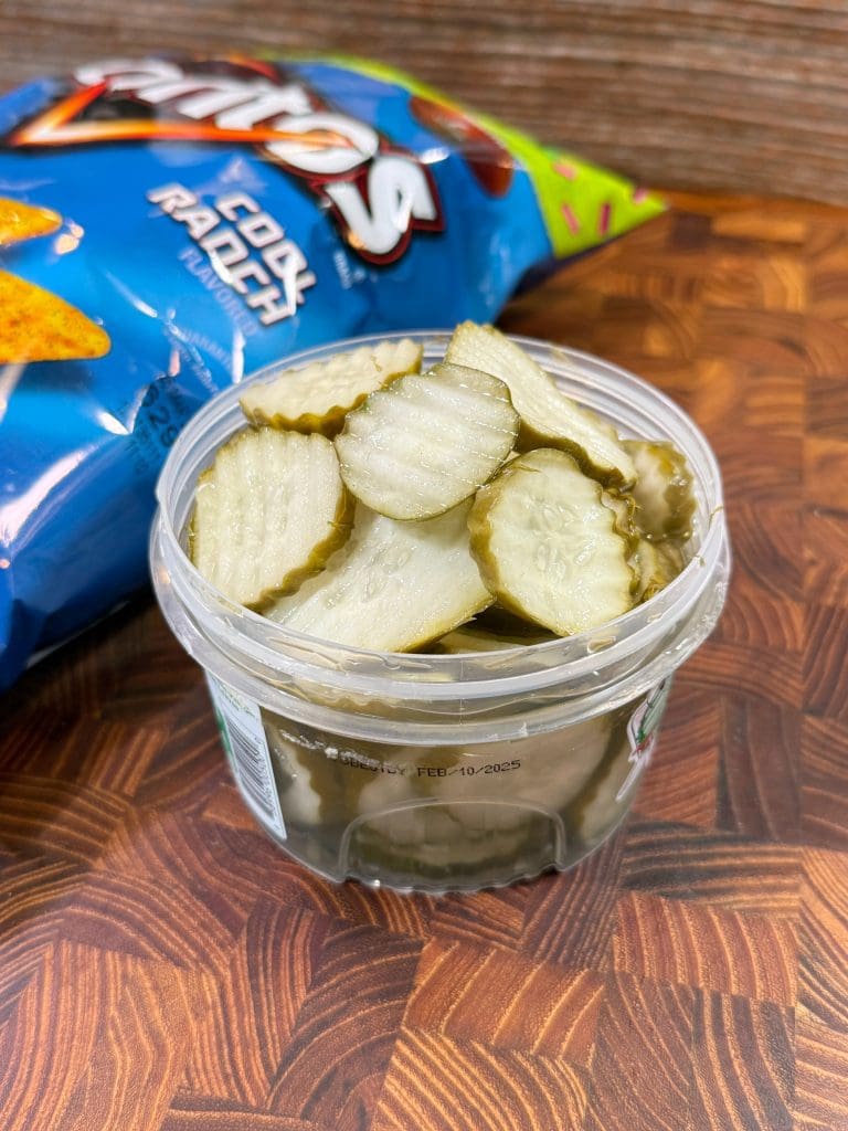 A clear plastic container filled with sliced pickles is on a wooden surface. Behind it is a partially visible bag of Doritos Cool Ranch chips, with part of the blue packaging and logo showing.