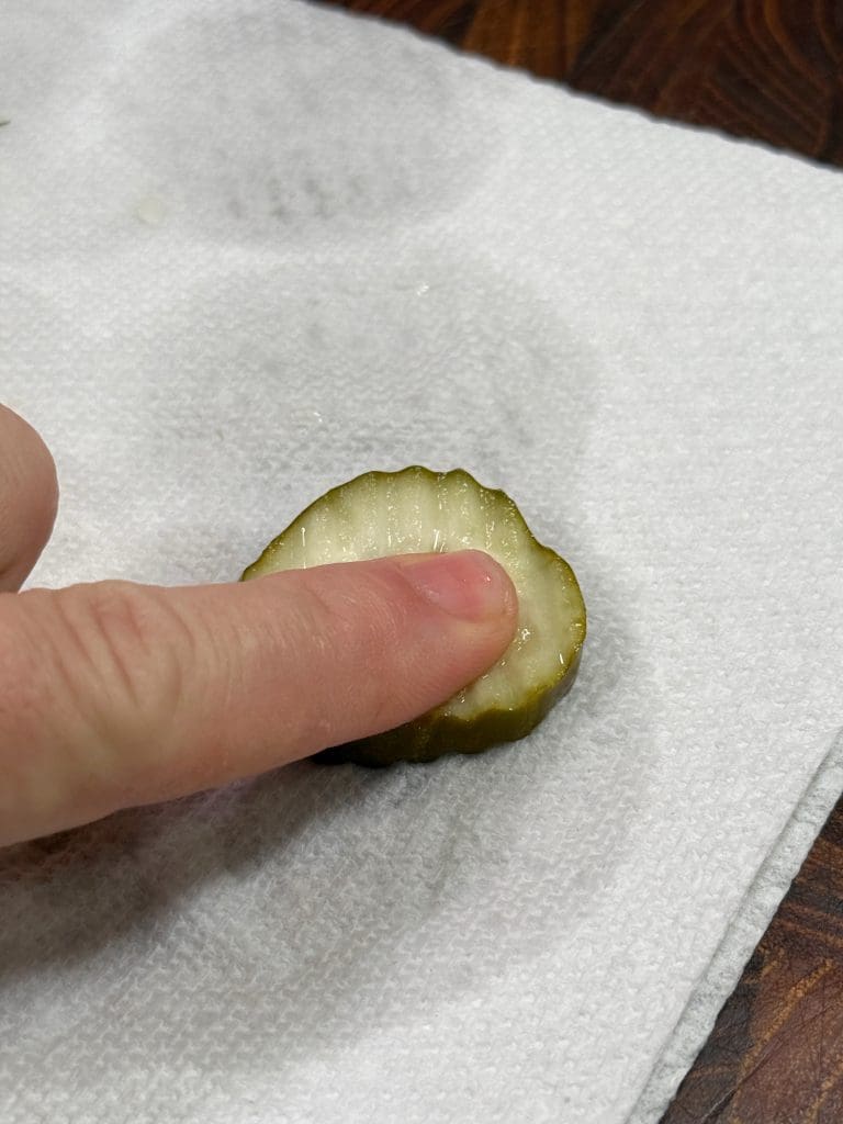 A hand with an extended finger pressing down on a slice of pickle that is placed on a white paper towel. The pickle slice appears moist, and the background is a wooden surface.