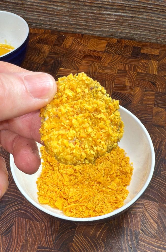A hand holding a round, breaded piece of food above a bowl filled with crushed, golden-brown crumbs. The countertop is wooden with a wavy pattern, and a small blue bowl is visible in the background.