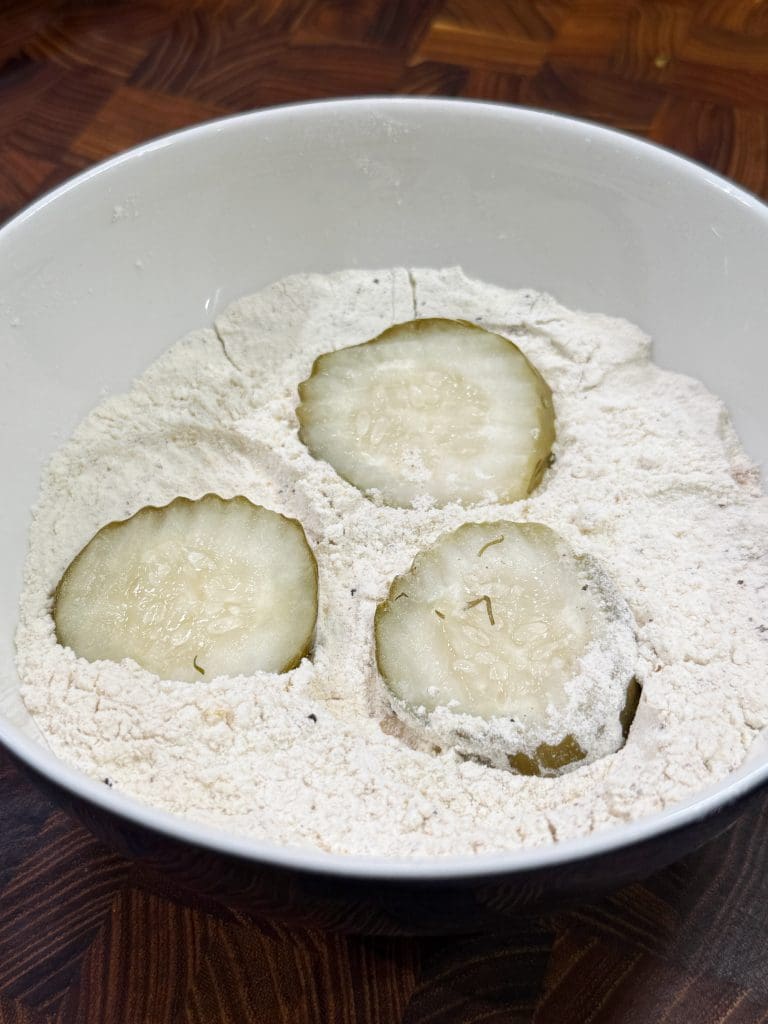 Three pickle slices sit in a white bowl of flour, ready for coating. The mixture rests on a wooden countertop, suggesting preparation for frying or another cooking method.
