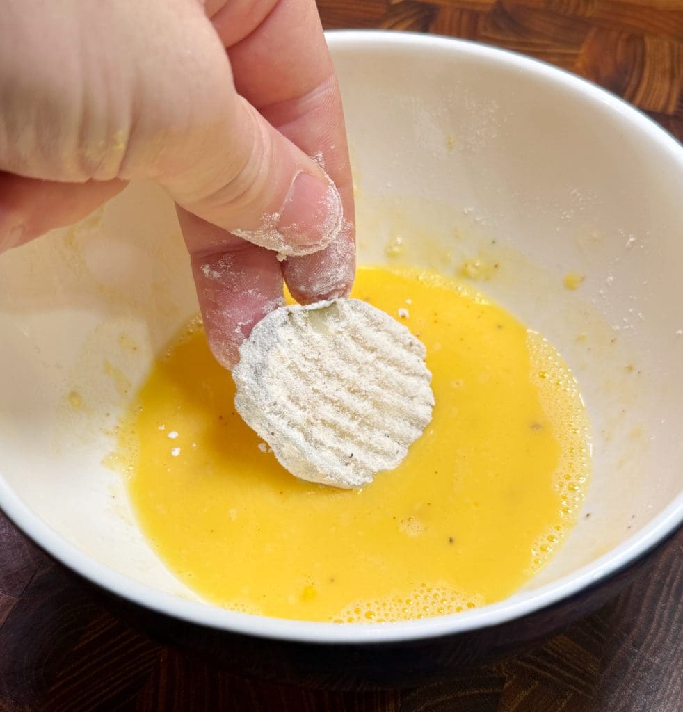 A hand dips a flour-coated slice into a bowl of beaten eggs. The bowl is on a wooden surface.