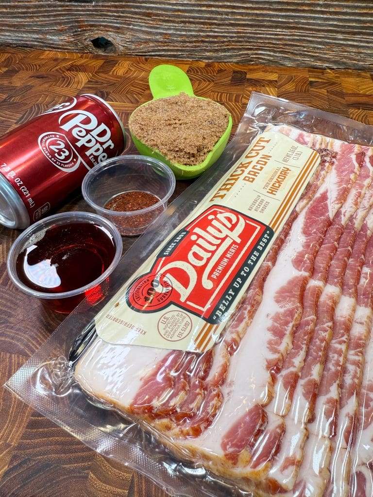 Package of sliced bacon on a wooden surface, surrounded by a can of Dr Pepper, a small bowl of red liquid, a cup of brown sugar, and a container with seasoning. A rustic wooden background completes the setup.