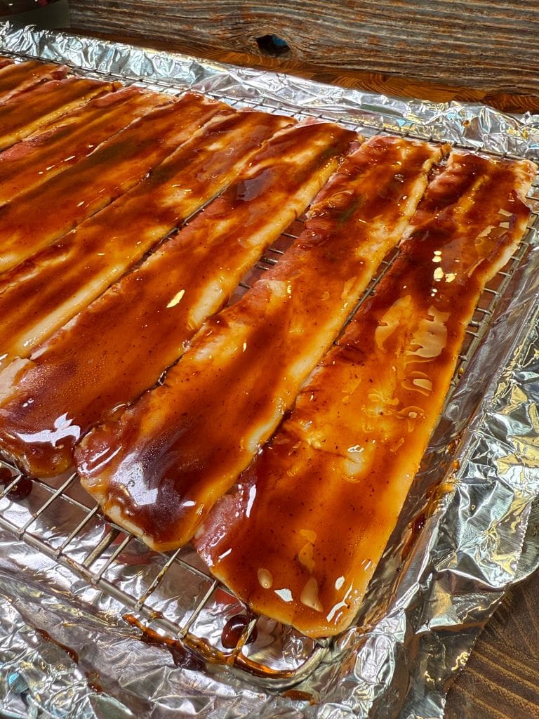 Slices of uncooked bacon arranged on a wire rack, coated with a glossy brown marinade, placed over foil-lined baking sheets. The background features a wooden surface.