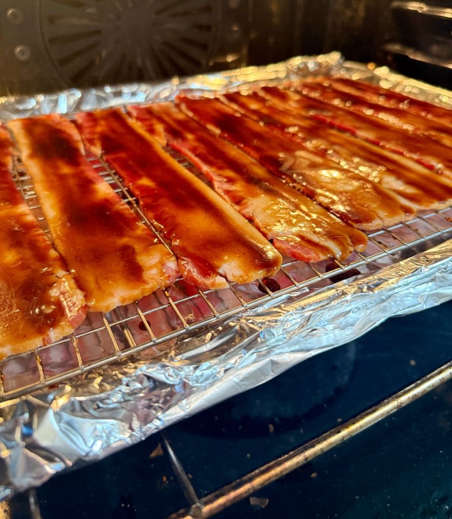 Slices of bacon coated in a brown glaze are arranged on a wire rack over a foil-lined baking sheet, placed inside an oven.