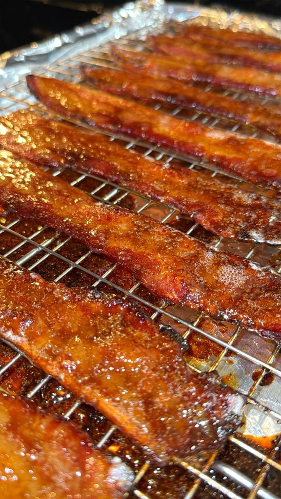 Close-up of crispy, glazed bacon strips resting on a wire rack over a foil-lined baking tray. The bacon is evenly spaced, with a shiny texture, indicating a sweet or savory coating.