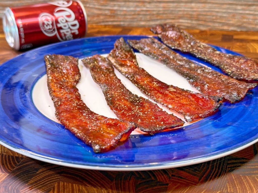 Five strips of crispy candied bacon on a blue and white plate, placed on a wooden surface. A can of Dr Pepper is visible in the background.