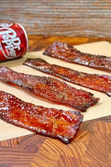 Four strips of candied bacon lay on a piece of parchment paper atop a wooden surface. A can of Dr Pepper is positioned beside the bacon. The bacon has a glossy, caramelized appearance.