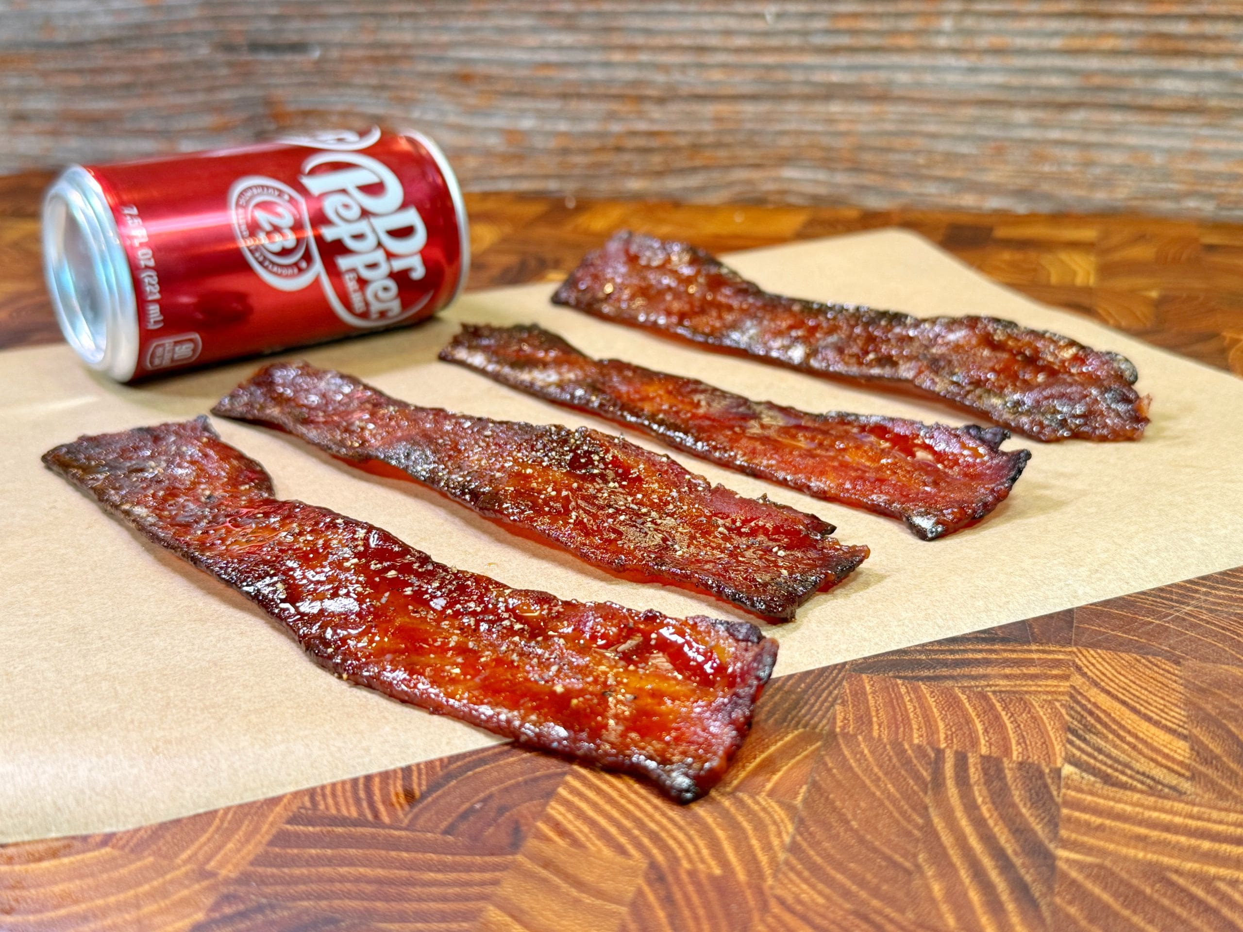 Four strips of candied bacon lay on a piece of parchment paper atop a wooden surface. A can of Dr Pepper is positioned beside the bacon. The bacon has a glossy, caramelized appearance.