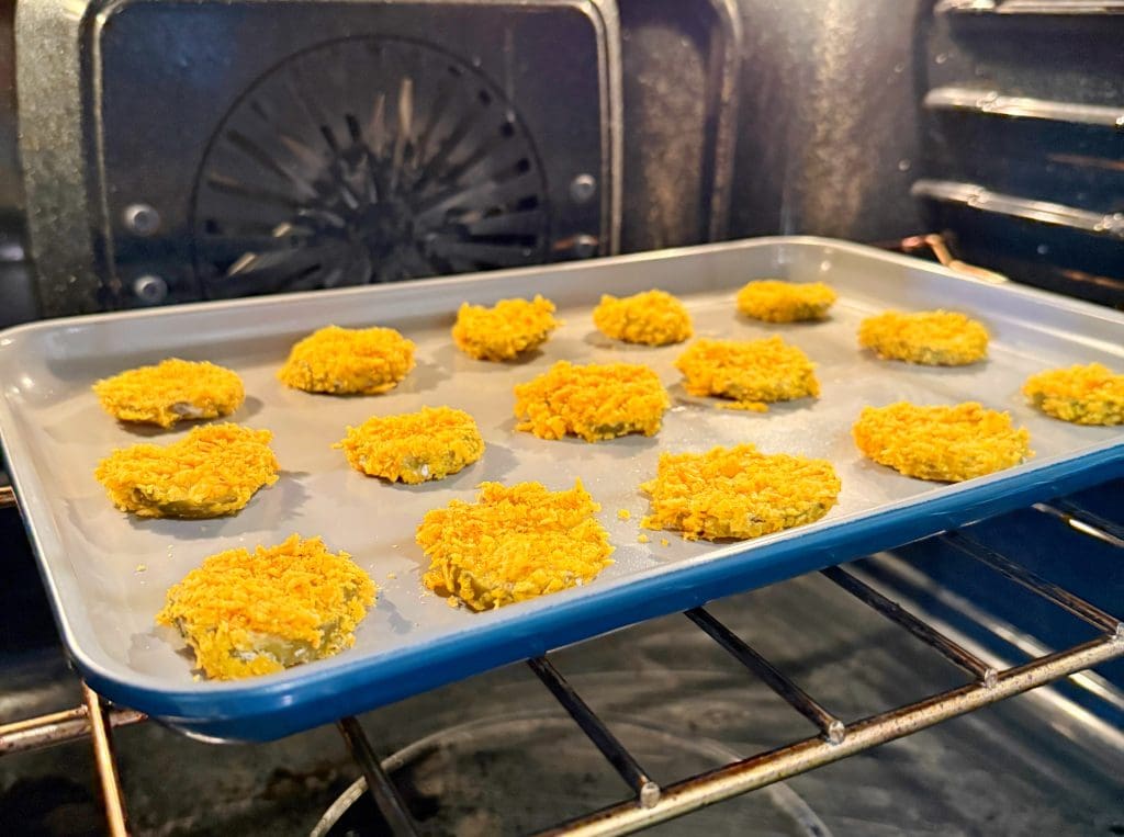 A baking sheet with rows of yellow-orange breaded items, possibly nuggets or patties, is placed on an oven rack. The oven interior is visible, with the back fan design evident. The items appear to be in the process of being baked.