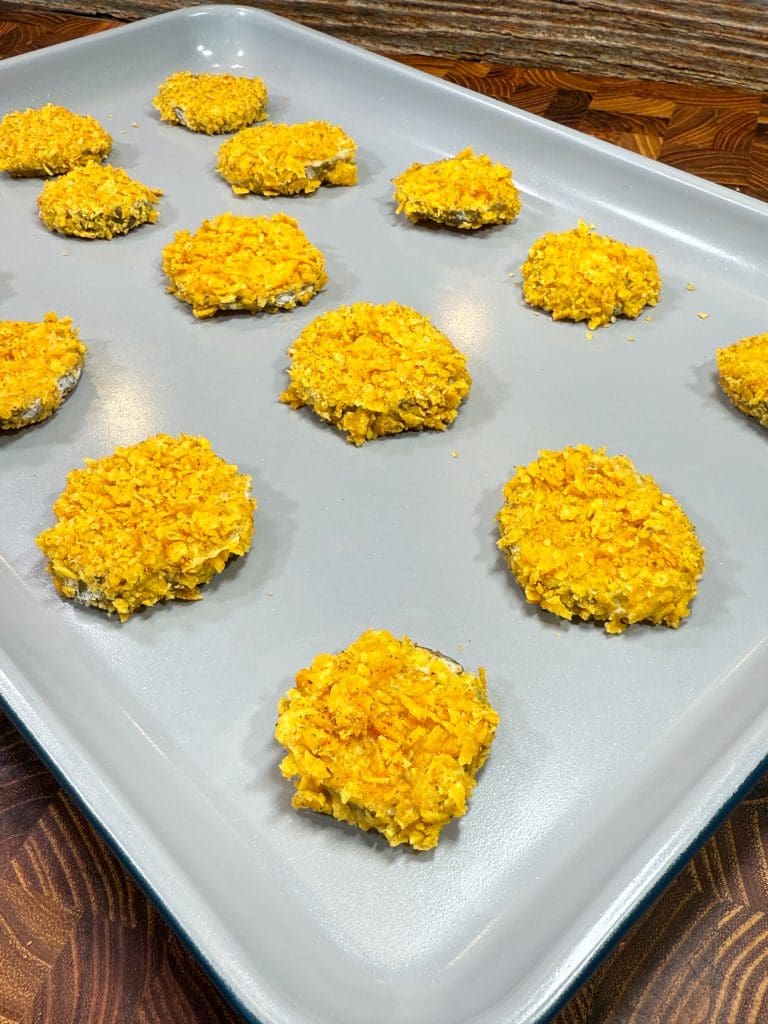 A baking tray with evenly spaced, round, breaded items ready to bake. They have a golden, crumbly coating and are set on a light gray baking sheet. The background shows a wooden surface.