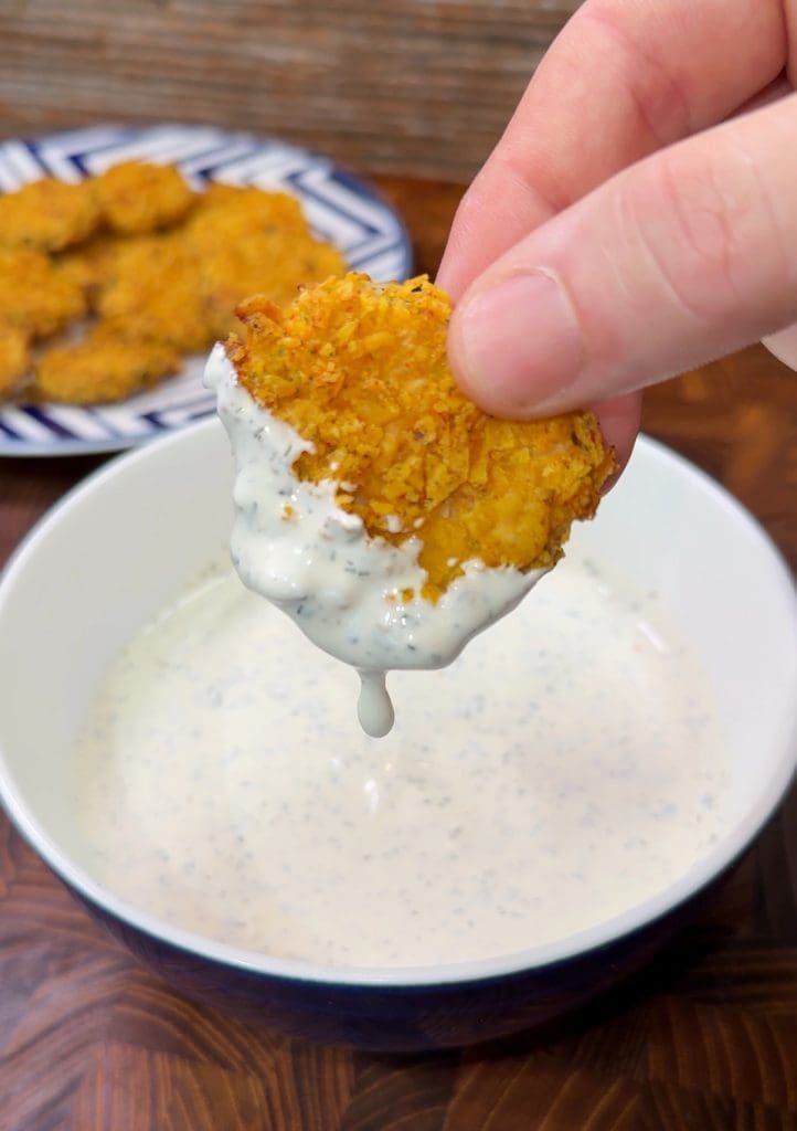 A hand holding a crispy, golden fried nugget dipped halfway into a white sauce inside a bowl. The nugget is coated with the sauce, and more nuggets can be seen on a plate in the background.