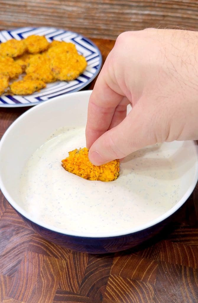 A hand dipping a crispy, orange-colored nugget into a bowl of creamy, speckled ranch dressing. A plate with more nuggets is visible in the background, set on a wooden surface.